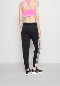 Rosa Sport-BH mit strukturierter Oberfläche und Racerback-Design, kombiniert mit schwarzen Hosen mit weißen Streifen an den Seiten. Schwarze Sportschuhe.