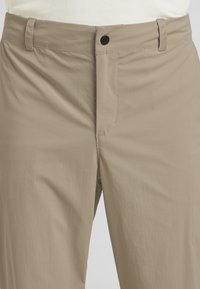Pantaloni in tessuto leggero beige, con una texture liscia, chiusura con bottone e passanti per cintura. Presentano cuciture minime e tasche laterali.