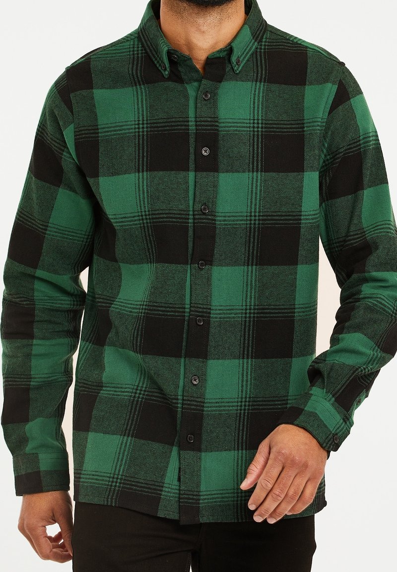 Homme portant une chemise à carreaux verte et noire à manches longues boutonnée avec col, visible du cou jusqu’aux hanches.