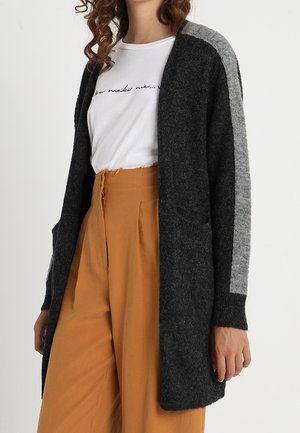 Kvinde iført en mørkegrå lang cardigan med en lysegrå stribe på ærmerne, hvid t-shirt med grafik og højtaljede sennepsfarvede bukser.