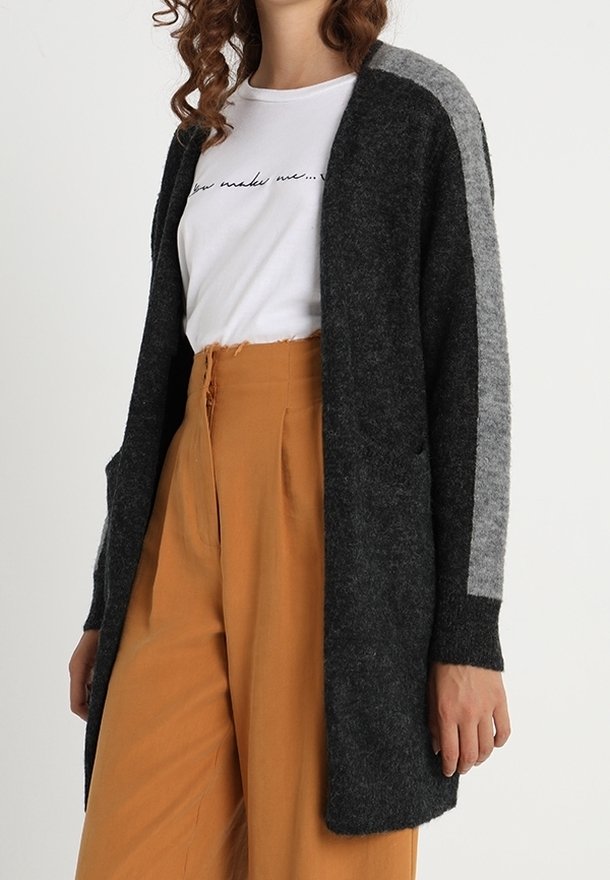 Femme portant un long cardigan gris foncé avec une bande grise claire sur les manches, un t-shirt blanc à motif et un pantalon taille haute moutarde.