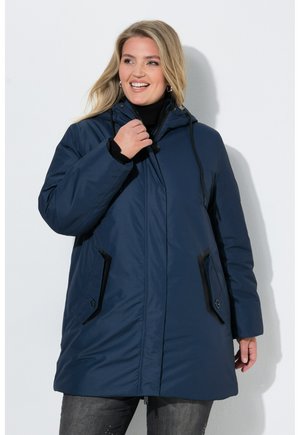 Femme souriante portant un manteau d'hiver bleu marine à capuche avec rabats de poche noirs et fermeture éclair noire, debout sur un fond blanc uni.