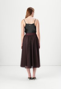 Een vrouw met lang bruin haar draagt een zwarte tanktop en een vloeiende burgundy tulle rok, staande op een witte achtergrond.