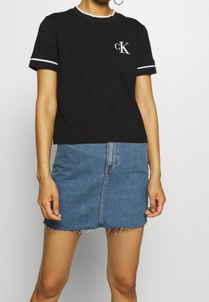 Czarny krótki t-shirt z białą lamówką i logo CK po lewej stronie klatki piersiowej, zestawiony z niebieską spódnicą jeansową z postrzępionym dołem i przednim szwem.