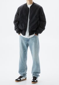 Blouson aviateur en nylon noir avec poignets côtelés, fermeture éclair à l'avant et poche latérale, associé à un jean ample bleu clair et des baskets noires.