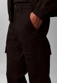 Személy fekete cargo nadrágot visel, kezét az oldalzsebben tartja, gombdíszítéssel és dzseki mandzsettájával szürke háttéren.