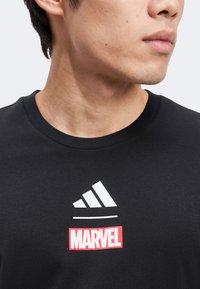 Νέος άνδρας που φορά μαύρο Τ-shirt με λευκό λογότυπο Adidas και κόκκινο λογότυπο Marvel στο στήθος, κοιτάζοντας προς τα δεξιά σε λευκό φόντο.