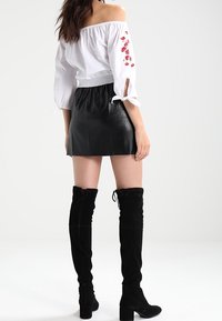 Blusa blanca sin mangas con bordado floral, combinada con una falda corta de cuero negra y botas de ante negras hasta el muslo.