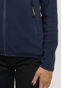 Giacca in pile blu navy con zip completa, superficie testurizzata e tasca laterale con zip. Pantaloni neri visibili sotto la giacca.