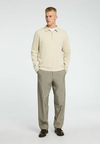 Suéter beige claro con cremallera y cuello plano, combinado con pantalones gris texturizados y zapatos marrones. Modelo de pie con las manos en los bolsillos.