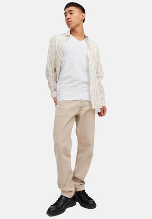 Ung mand iført beige stribet skjorte over hvid T-shirt, beige bukser og sorte snørebåndssko, stående mod en hvid baggrund.