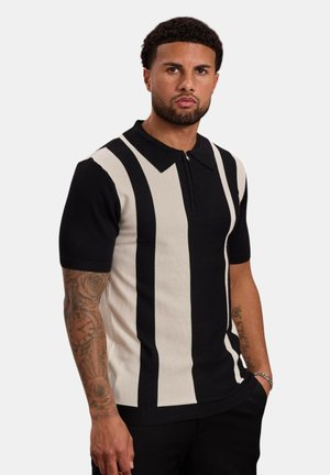 Homme aux cheveux courts et bouclés avec une barbe, portant un polo à manches courtes rayé verticalement noir et blanc et un pantalon noir, avec des tatouages sur les bras.