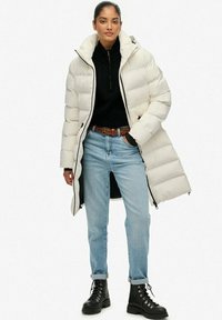 Gewatteerde jas in off-white met een hoge kraag, ritssluiting en doorgestikte textuur. Zwarte trui eronder, lichtblauwe spijkerbroek en zwarte veterlaarzen.