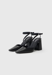 Musta nahkainen slingback-korkokenkä, jossa on teräväkärkinen muoto, säädettävä nilkkaremmi ja neliömäinen korko, jonka pinnassa on sileä tekstuuri ja minimalistinen muotoilu.