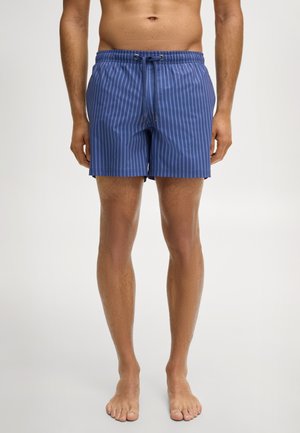 Mann trägt blaue, vertikal gestreifte Badeshorts mit elastischem Bund und Kordelzug, steht barfuß vor einem einfachen Hintergrund.