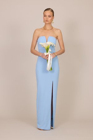 Γυναίκα με ανοιχτό μπλε φόρεμα χωρίς τιράντες κρατά ένα μπουκέτο λευκές κάλλες με άσπρη κορδέλα, στέκεται μπροστά σε μπεζ φόντο.