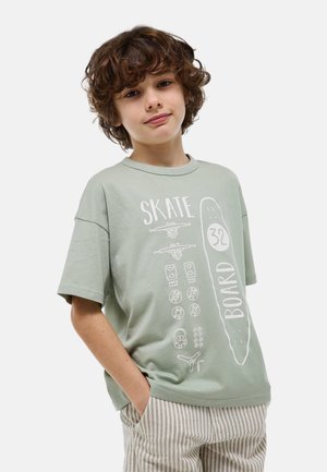 Garçon aux cheveux bouclés bruns portant un t-shirt vert avec un imprimé de pièces de skateboard et un pantalon beige rayé, debout les mains dans les poches.