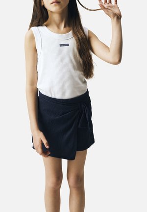 Fille aux longs cheveux bruns portant un haut blanc sans manches côtelé et une jupe portefeuille bleu marine, tenant une mèche de cheveux d'une main.