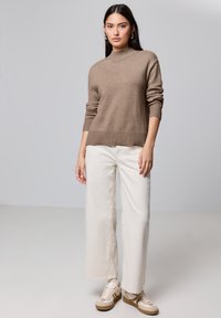 Hellbrauner Strickpullover mit hohem Kragen und Rippbündchen, kombiniert mit beigen, weit geschnittenen Jeans und cremefarbenen Sneakern. Einfaches Design, keine Muster.