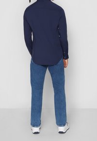 Marinblå skjorta med knappar, långa ärmar och rundad nederkant, tillsammans med blå jeans. Har en slät textur och standard kragdesign.