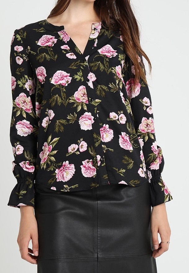 Femme portant un chemisier noir à imprimé floral rose et manches longues, associé à une jupe en cuir noire.