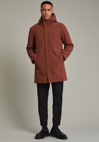 Mantou maro, căptușit, cu glugă, închidere cu fermoar frontal și buzunare laterale. Purtați peste pantaloni negri și pantofi, evidențiind un design elegant.
