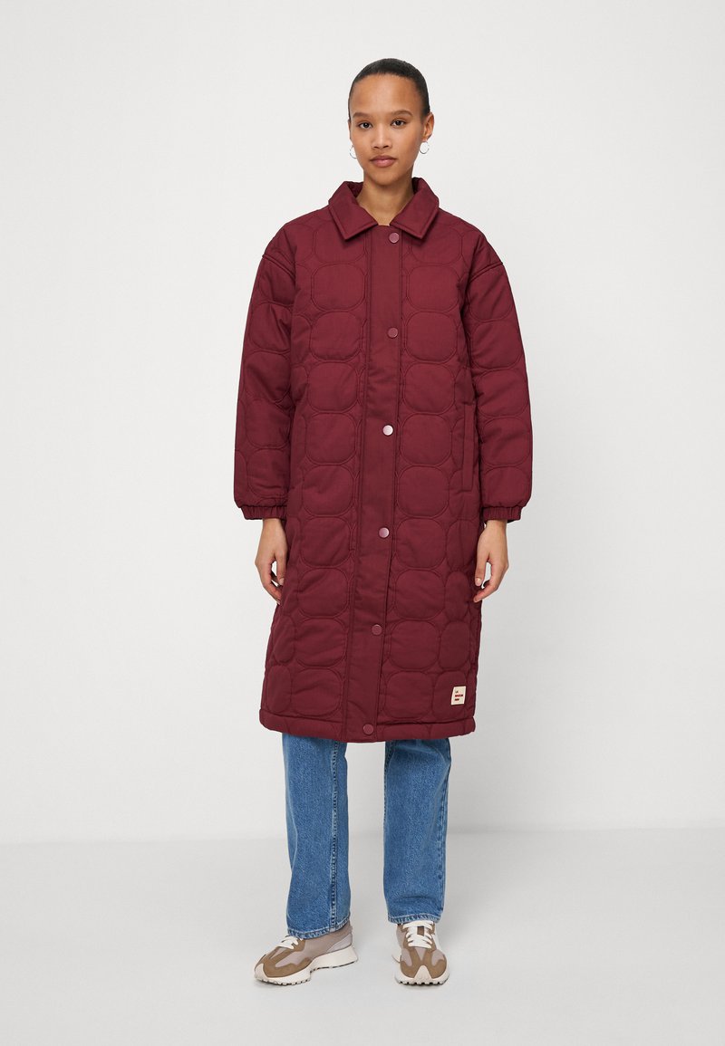 Manteau long rembourré bordeaux avec un devant à boutons, un col rond et un design matelassé. Porté sur un jean bleu et des baskets claires. Fond uni.