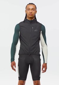 Young man with braids wearing black cycling vest over green and white long-sleeve top and black padded cycling shorts, standing facing forward.