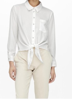Kobieta nosząca białą koszulę na guziki, zawiązaną w pasie, z przednią kieszenią, w połączeniu z beżowymi spodniami na białym tle.