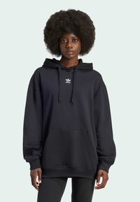 Svart oversized hettegenser laget av bomullsmiks, med en frontlomme, snøring i hetten, og hvit Adidas-logo på brystet.