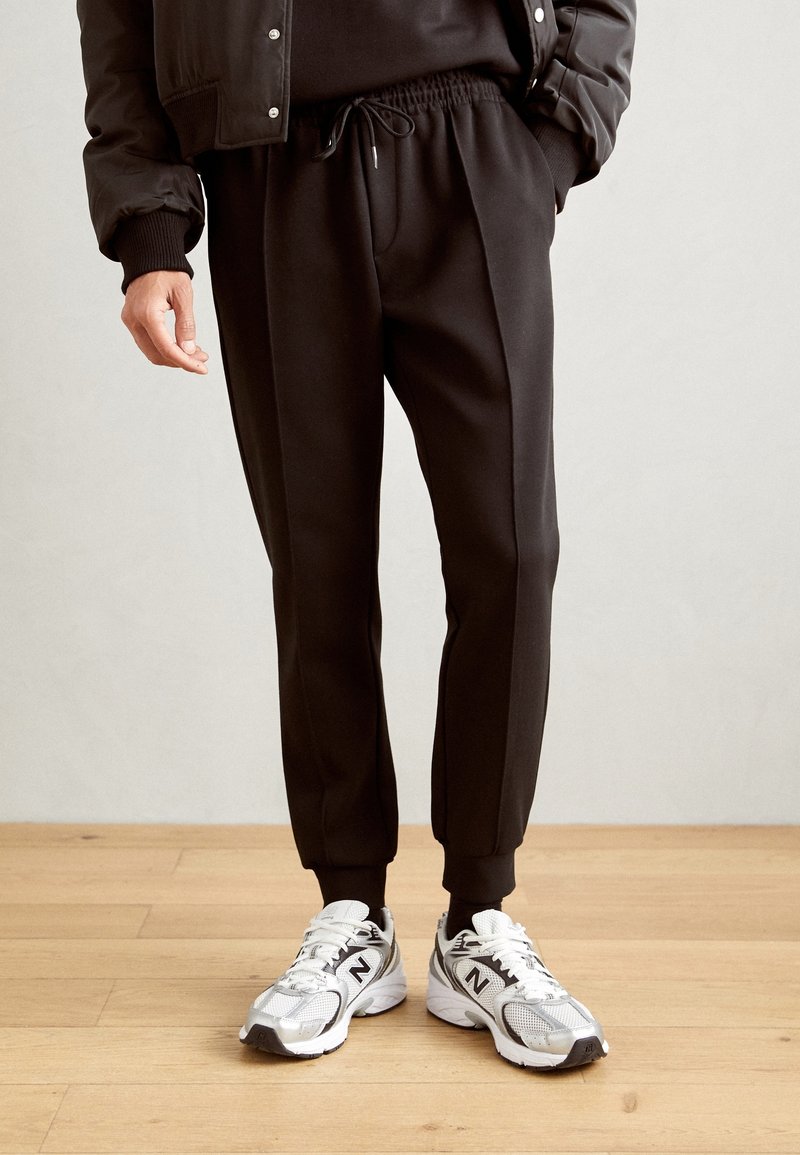 Svarta joggers med dragsko i midjan och ribbade muddar, ihop med grå och vita träningsskor med mesh-överkupper och logodetaljer.