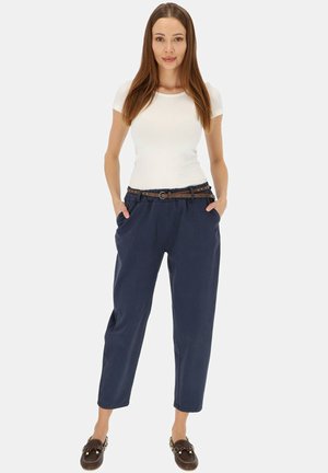 Kobieta stoi z rękami w kieszeniach, ubrana w biały krótki rękaw, granatowe spodnie z brązowym paskiem oraz brązowe mokasyny.