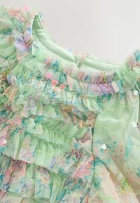 Zöld ruha, amely puha tüll rétegezett fodrokkal és pasztell virágmintákkal díszített. Kerek nyakkivágás, apró flitterekkel díszítve.