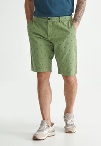 Shorts en coton verts avec un motif d'ancre, deux poches latérales et une fermeture par bouton, associés à des baskets beige et gris.