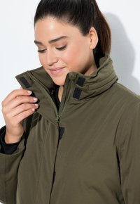 Mujer con cabello oscuro abrochando el cuello de su chaqueta verde con cremallera y cierres de Velcro.