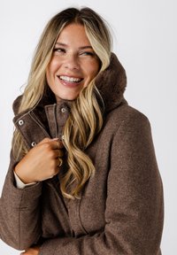 Bruine wollen mix jas met een bontachtige capuchon, voorzien van een ritssluiting en drukknoopsluiting, en een zachte textuur. Het model lacht terwijl ze de kraag vasthoudt.