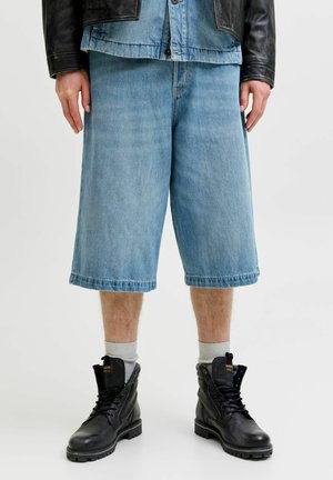 Mann trägt hellblaue knielange Denim-Shorts, schwarze Lederstiefel mit weißen Socken und eine schwarze Lederjacke über einem Denimhemd.