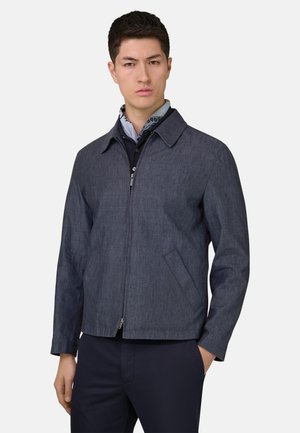 Mann trägt dunkle blaue Reißverschlussjacke über marineblauem Hemd und gemustertem Halstuch, mit einer Hand in der Tasche, neutraler Hintergrund.