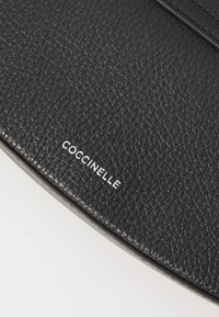 Borsa a mano in pelle nera con finitura testurizzata, forma rotonda e marchio in argento "Coccinelle" stampato sulla parte anteriore.