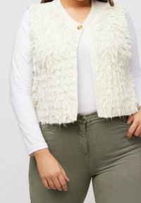 Strukturiertes weißes Strickweste mit Fransen-Design, ausgestattet mit einem einzelnen Knopfverschluss, getragen über einem weißen Langarmshirt und olivgrünen Hosen.