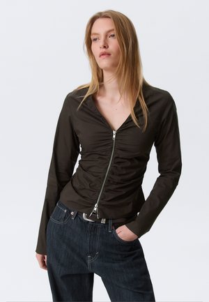 Σκούρο ελιάς πουκάμισο με φερμουάρ, με λεπτομέρειες ραφής, μακριά μανίκια και ντεκολτέ V, συνδυασμένο με εφαρμοστά σκούρα τζιν.