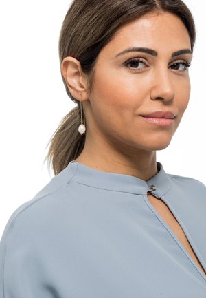 Hängende Ohrringe mit einer goldenen Kette und einem weißen Perlenakzent. Die Bluse ist hellblau, hat einen Stehkragen und einen kleinen Verschluss.