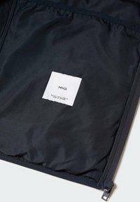 Intérieur de veste noire avec une étiquette de marque rectangulaire blanche MNG et des détails de taille et de fabrication sur un tissu noir près de la fermeture éclair.