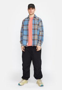 Blauw en oranje geruite flanellen shirt over een koraalkleurig T-shirt, gecombineerd met zwarte cargobroeken en multicolore sneakers, met een zwart petje.