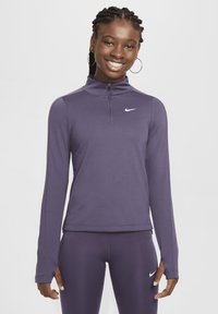Joven sonriente, vistiendo una chaqueta con cremallera deportiva negra de Nike y leggins morados a juego con logotipos de swoosh blancos.