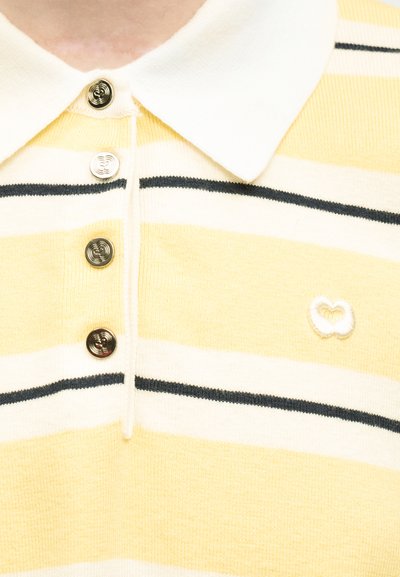 Κοντινό πλάνο κίτρινης και λευκής ριγωτής μπλούζας πόλο με μαύρες ρίγες, λευκό γιακά, τέσσερα μεταλλικά κουμπιά και ένα μικρό κεντητό λογότυπο.