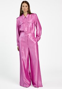 Vrouw met lang haar, gekleed in een glanzende roze blouse met knopen en een wijdere broek, staand met één hand in de zak tegen een witte achtergrond.