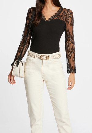 Femme portant un haut en dentelle noire à manches longues, un pantalon clair taille haute avec une ceinture blanche, et portant un petit sac à épaule blanc.