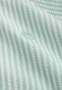 Světlé zelené plátno s pravidelnými vertikálními pruhy, které střídají strukturované a hladké části, doplněné malým vyšívaným motivem slunce ve spodní části.