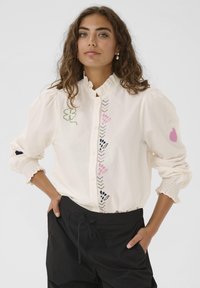 Donna con lunghi capelli ricci castani, che indossa una camicia bianca con motivi ricamati colorati e pantaloni neri, in posa con le mani nelle tasche.